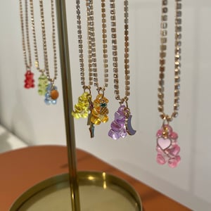 May include: A display of gold chains with colorful gummy bear charms. The charms include a yellow bear with a pineapple, a purple bear with a crescent moon, and a pink bear with a heart.