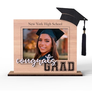 May include: A wooden picture frame with a graduation cap on top. The frame has a photo of a graduate in a cap and gown. The text "New York High School" and "Congrats Grad" is on the frame.
