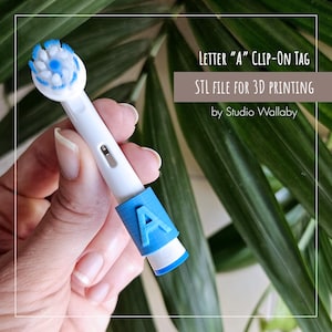 Puede incluir: Un cabezal de cepillo de dientes blanco y azul con una etiqueta azul con la letra "A". El texto en la imagen dice "LETTER 'A' CLIP-ON TAG" y "STL FILE FOR 3D PRINTING".