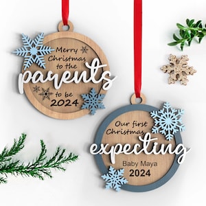 Op de afbeelding: Twee houten kerstversieringen met blauwe sneeuwvlokken. De eerste versiering zegt "Merry Christmas to the parents to be 2024". De tweede versiering zegt "Our first Christmas expecting Baby Maya 2024".