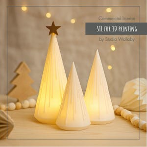 May include: Three white cone-shaped Christmas trees with vertical lines and a warm glow. The tallest tree is topped with a wooden star. The text "STL FOR 3D PRINTING" is visible. A wooden Christmas tree and string of beads are in the background.