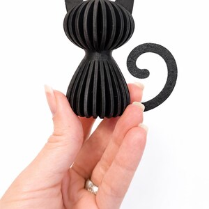 Puede incluir: Figura de gato negro con un dise&ntilde;o acanalado &uacute;nico. El gato tiene una forma estilizada con una cola curvada y orejas puntiagudas. La figura est&aacute; hecha de un material negro s&oacute;lido y se sostiene en una mano sobre un fondo blanco.