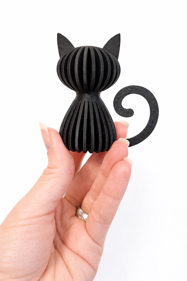 Puede incluir: Figura de gato negro con un dise&ntilde;o acanalado &uacute;nico. El gato tiene una forma estilizada con una cola curvada y orejas puntiagudas. La figura est&aacute; hecha de un material negro s&oacute;lido y se sostiene en una mano sobre un fondo blanco.