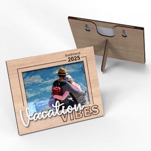 May include: A wooden photo frame with a picture of a couple in front of a mountain landscape. The frame has the text "Vacation Vibes" and "Swithland 2025" printed on it.