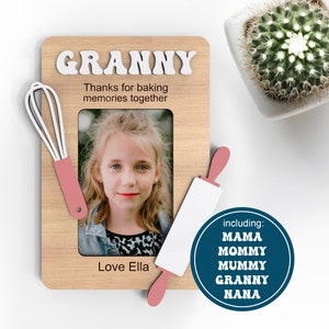 May include: Wooden photo frame with a picture of a young girl, a pink rolling pin, and a pink whisk. The frame has the text "GRANNY" and "Thanks for baking memories together". The text "Love Ella" is at the bottom of the frame. The text "including: MAMA MOMMY MUMMY GRANNY NANA" is in a blue circle.
