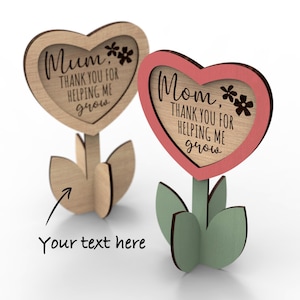 Op de afbeelding: Twee houten hartvormige bloemenversieringen met de tekst "Mum, Thank you for helping me grow" en "Mom, Thank you for helping me grow". De bloemen zijn roze en bruin met groene bladeren. De tekst "Your text here" staat onder de bloemen.