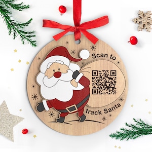 May include: A round wooden bauble featuring a cartoon image of Santa Claus carrying a sack with a QR code. The bauble has a red ribbon and the text "Scan to track Santa".