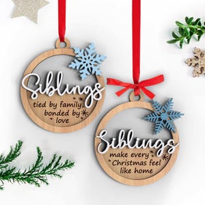 May include: Two wooden Christmas ornaments with the word "Siblings" in white script. Each ornament has a light blue snowflake and is suspended by a red ribbon. One ornament reads "tied by family, bonded by love". The other reads "make every Christmas feel like home".