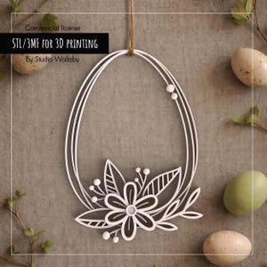 May include: White, laser-cut Easter egg decoration with a floral design. The egg-shaped frame is suspended by a twine string. The text "STL/3MF FOR 3D PRINTING" is displayed on a black rectangle. Decorative eggs and greenery are in the background.