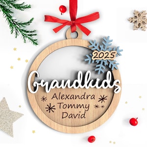 May include: Wooden Christmas ornament with the word "Grandkids" in white script. The ornament has a light brown wooden frame with the names Alexandra, Tommy, and David. A blue snowflake with the year 2025 is attached.