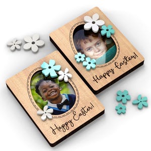 May include: Two wooden photo frames with oval cutouts, each holding a child's photo. The frames are decorated with white and light blue flower accents and the words "Happy Easter!" in script. Additional flower embellishments are scattered around.