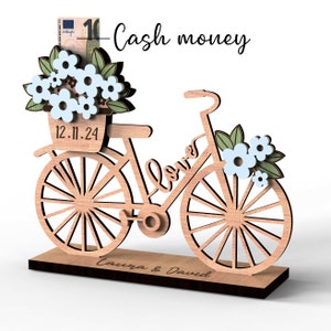 May include: Wooden bicycle money box with a basket of flowers and the text "Cash money" and "Love". The base of the box is engraved with the date "12.11.24" and the names "Laura & David".