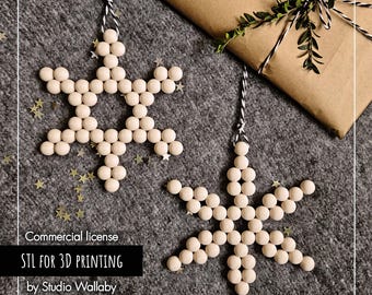 Puede incluir: Dos adornos de copos de nieve de madera hechos de pequeñas cuentas redondas, colgados de hilo blanco y negro. Los copos de nieve están sobre una superficie gris con un regalo envuelto y el texto "STL FOR 3D PRINTING by Studio Wallaby."