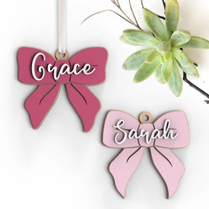 May include: Two wooden bow-shaped ornaments in shades of pink. The top ornament is a deeper pink with the name "Grace" in white script. The bottom ornament is a lighter pink with the name "Sarah" in white script. A white ribbon is attached to the top ornament.