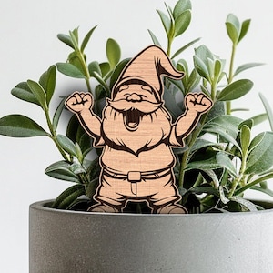 May include: Wooden gnome figurine with a cheerful expression, arms raised in the air, wearing a hat and trousers. The gnome is standing in a grey planter with green plants.