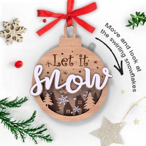 May include: Wooden Christmas ornament shaped like a ball with the text "Let it snow" in white lettering. The ornament has a cut-out design of a winter scene with trees, houses, and snowflakes.