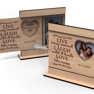 Op de afbeelding: Een houten fotolijst met een hartvormige uitsparing voor een foto. De lijst heeft een citaat dat luidt "Leef elk moment, Lach tot het pijn doet, Liefheb als een gek." De datum "02.14.2024" is ook gegraveerd op de lijst.