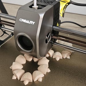 May include: A gray Creality 3D printer extruder head is positioned over a build plate, actively printing multiple small, light beige, mushroom-shaped objects arranged in a circular pattern. A white filament tube enters the top of the print head.