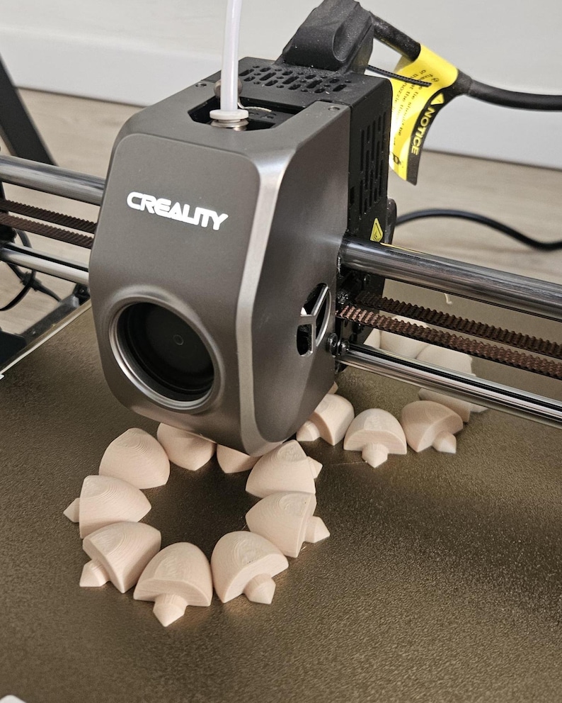 May include: A gray Creality 3D printer extruder head is positioned over a build plate, actively printing multiple small, light beige, mushroom-shaped objects arranged in a circular pattern. A white filament tube enters the top of the print head.