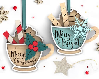 Könnte beinhalten: Zwei hölzerne Weihnachtsornamente in Form von Tassen mit heißer Schokolade. Eine Tasse hat einen blauen Hintergrund mit dem Text "Merry & Bright" und einer Schneeflocke. Die andere Tasse hat einen braunen Hintergrund mit dem Text "Merry Christmas!" und einem Zweig mit Stechpalme und roten Beeren.