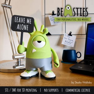 Pode incluir: Uma figura de monstro verde-limão com um olho e dois pequenos chifres, vestindo calções cinzentos e ténis brancos. A figura segura uma placa retangular preta com o texto branco LEAVE ME ALONE. A personagem tem um dente e uma expressão aborrecida.