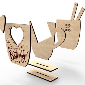 May include: Wooden craft kit to make a mug shaped birthday card. The card features a heart-shaped cutout and the text "Happy Birthday!" and "Hug in a Mug".