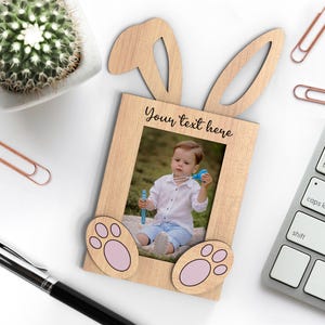 Op de afbeelding: Houten fotolijst met een konijnenthema, met oren en pootafdrukken. De lijst bevat een foto van een kind. De tekst "Your text here" staat boven de foto. Een schattig paascadeau.
