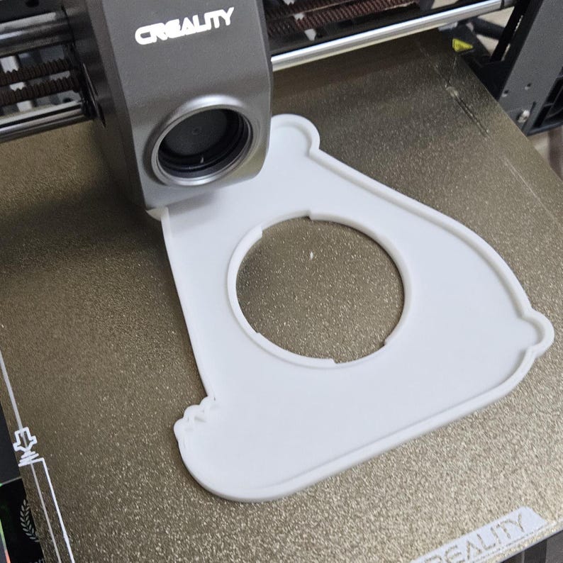 May include: A white, 3D-printed object with a circular cutout, positioned on a 3D printer bed. The printer head, labeled "CREALITY," is visible above. The object's design features rounded edges and a unique shape, suggesting a custom-made part.