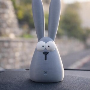 May include: A light gray, cartoon-style bunny figurine with large, expressive eyes and long ears. The bunny has a simple, minimalist design with a small black nose and a cross-shaped mouth. The figurine is sitting on a dark surface.