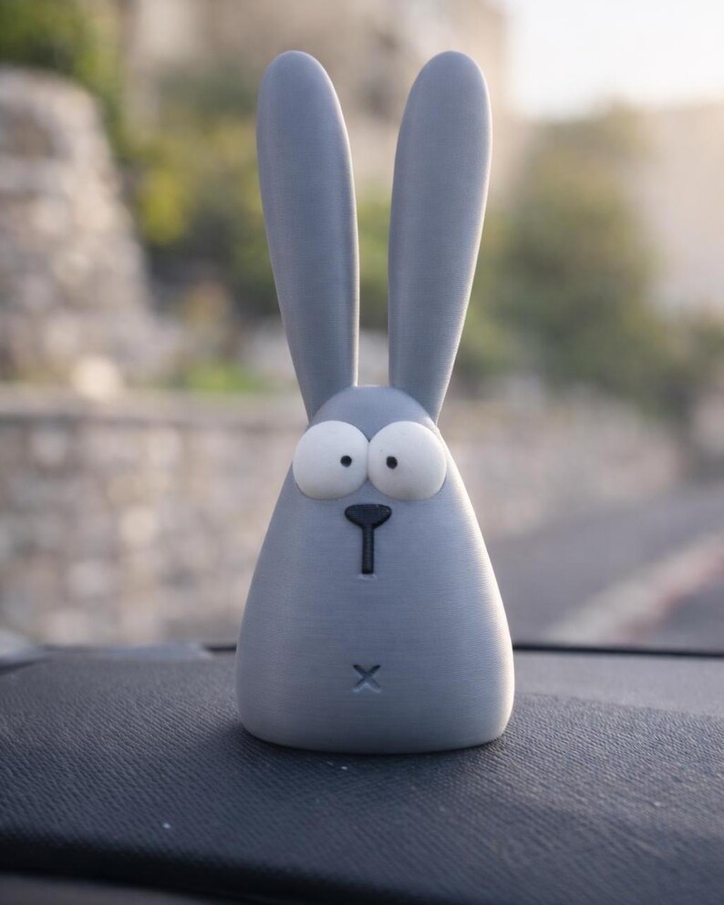 May include: A light gray, cartoon-style bunny figurine with large, expressive eyes and long ears. The bunny has a simple, minimalist design with a small black nose and a cross-shaped mouth. The figurine is sitting on a dark surface.