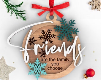 May include: Wooden Christmas ornament shaped like a circle with the words "Friends are the family you choose" written on it. The ornament is decorated with snowflakes in shades of blue, green, and brown.