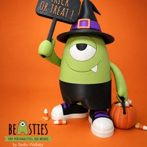 May include: A green one-eyed monster figurine wearing a black witch hat with a purple band and black shorts. It holds a black sign that reads TRICK OR TREAT! and carries an orange pumpkin bucket filled with candy corn on a bright orange background.