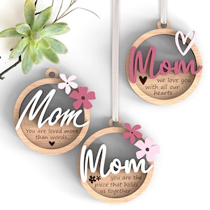 Peut inclure: Trois ornements en bois pour la fête des mères avec des accents floraux et des rubans blancs pour suspendre. Chaque ornement présente le mot "Mom" en écriture cursive, ainsi qu'un message sincère. Les ornements sont décorés de fleurs roses et blanches.