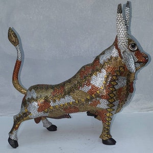 May include: A wooden bull figurine with a geometric pattern in shades of gold, brown, and white. The bull is standing on all four legs with its head turned to the right.