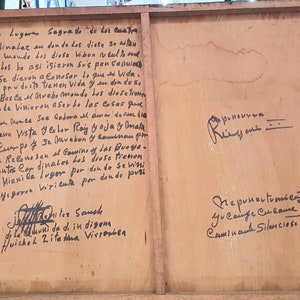 May include: A close-up of a wooden sign with handwritten text in black ink. The text is in Spanish and discusses the four cardinal directions and the gods. The sign is weathered and has a rustic appearance.