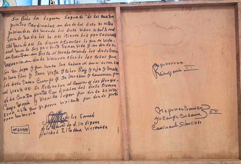 May include: A close-up of a wooden sign with handwritten text in black ink. The text is in Spanish and discusses the four cardinal directions and the gods. The sign is weathered and has a rustic appearance.