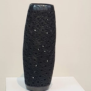Puede incluir: Un jarrón de cerámica negro con un diseño único e intrincado de patrones en espiral y agujeros recortados. El jarrón se muestra en un pedestal blanco.