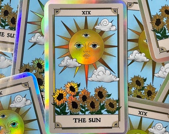 Puede incluir: Una carta de tarot holográfica con un borde dorado y un sol con una cara y un tercer ojo en el centro. La carta está rodeada de girasoles y nubes. El texto "XIX THE SUN" está impreso en la carta.