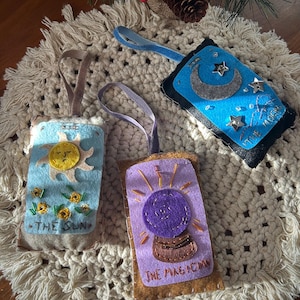 Könnte beinhalten: Drei handgefertigte Tarotkarten-Ornamente aus Filz. Das Sonnen-Ornament ist hellblau mit einer gelben Sonne und gestickten Blumen. Das Mond-Ornament ist blau mit einem silbernen Mond und Sternen. Das Magier-Ornament ist lila mit einer Kristallkugel.