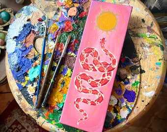 May include: A pink canvas painting featuring a white snake design. The snake is curled up and has a red and white pattern. The background is a light pink with a yellow sun in the top centre.