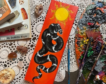 May include: A hand-painted canvas featuring a black and white snake on an orange background. The snake is curled up in a circle and has a white stripe down its back. The background is a bright orange with a yellow sun in the top right corner.
