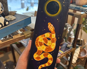 May include: A rectangular canvas painting depicting a yellow and orange snake on a dark blue backdrop, with a yellow-rimmed black circle and small yellow dots. The snake is coiled and detailed, with a textured appearance.