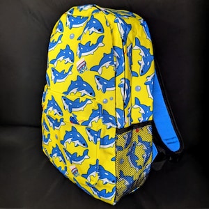 May include: A yellow backpack with a blue shark pattern. The backpack has a mesh pocket on the side and a blue zipper pull.