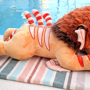 Puede incluir: Un peluche de le&oacute;n en posici&oacute;n tumbada, con cuerpo beige y detalles a rayas rojas y blancas. El le&oacute;n tiene melena marr&oacute;n y ojos rojos, descansando sobre una toalla a rayas cerca de una piscina.