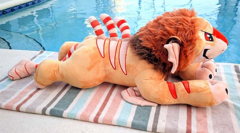 Puede incluir: Un peluche de le&oacute;n en posici&oacute;n tumbada, con cuerpo beige y detalles a rayas rojas y blancas. El le&oacute;n tiene melena marr&oacute;n y ojos rojos, descansando sobre una toalla a rayas cerca de una piscina.