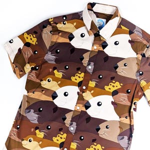 May include: A brown and white short-sleeved button-down shirt with a pattern of sea otters. The shirt has a brown background with white sea otters with black eyes and noses. The shirt has a button-up collar and a button placket.