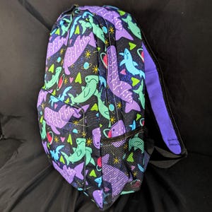 May include: A black backpack with a repeating pattern of purple and teal whale sharks, planets, and stars. The backpack has a mesh side pocket, a front pocket, and purple straps and trim. The bag is designed for carrying books and supplies.