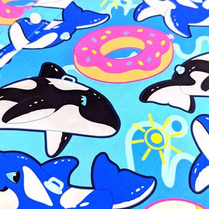 May include: A blue and white patterned fabric with a repeating design of orcas, donuts, and beach balls. The orcas are in different poses, some are swimming, some are jumping, and some are floating on their backs. The donuts are pink with yellow frosting and sprinkles. The beach balls are pink and white.