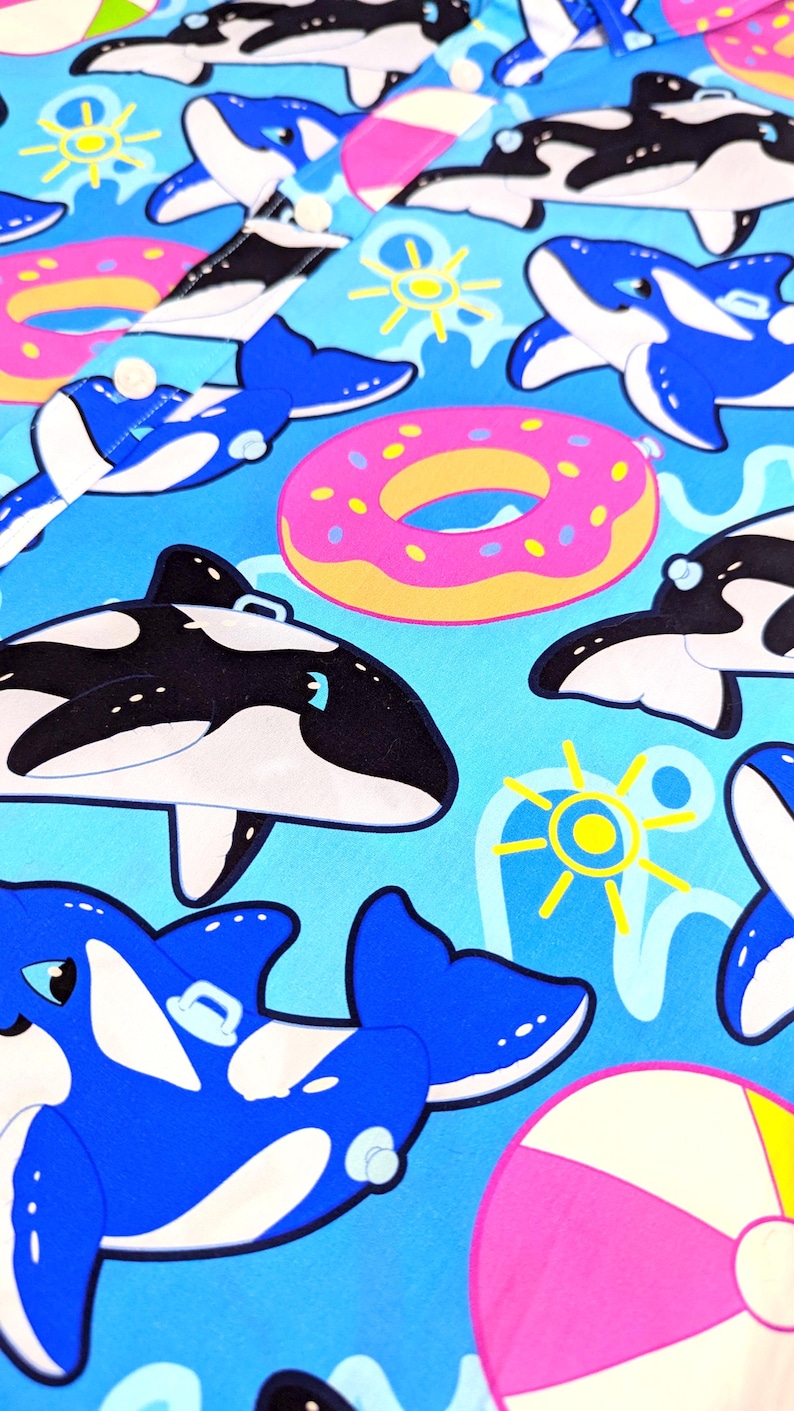 May include: A blue and white patterned fabric with a repeating design of orcas, donuts, and beach balls. The orcas are in different poses, some are swimming, some are jumping, and some are floating on their backs. The donuts are pink with yellow frosting and sprinkles. The beach balls are pink and white.