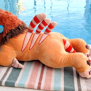 Puede incluir: Un juguete de peluche con forma de le&oacute;n, con pelaje naranja, melena marr&oacute;n y detalles a rayas rojas y blancas. El le&oacute;n tiene una etiqueta azul en la oreja y est&aacute; tumbado en una toalla a rayas cerca de una piscina.